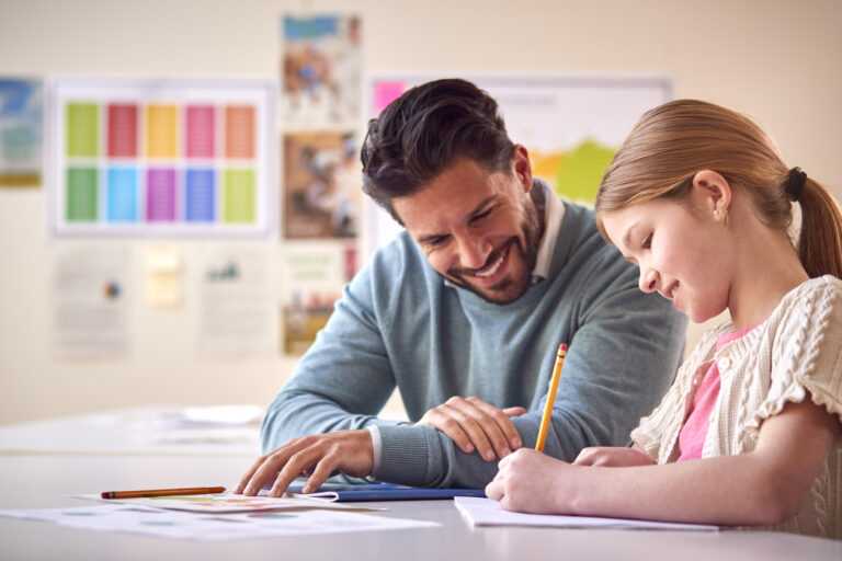 Male Teacher With Female Student In School Classroom Sitting At Desk Writing In Book Together